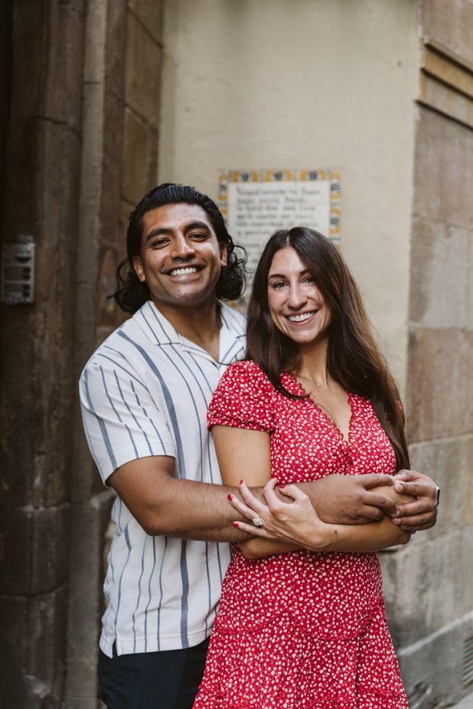 A smiling couple stands close together in an outdoor setting. The man wears a short-sleeved striped shirt and hugs the woman from behind. The woman wears a red patterned dress and smiles at the camera.