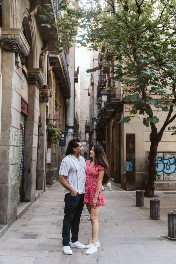 A couple stands smiling at each other on a narrow, cobblestone street lined with old buildings and trees. The man wears a striped shirt and dark pants, and the woman wears a pink dress. It appears to be a sunny day.