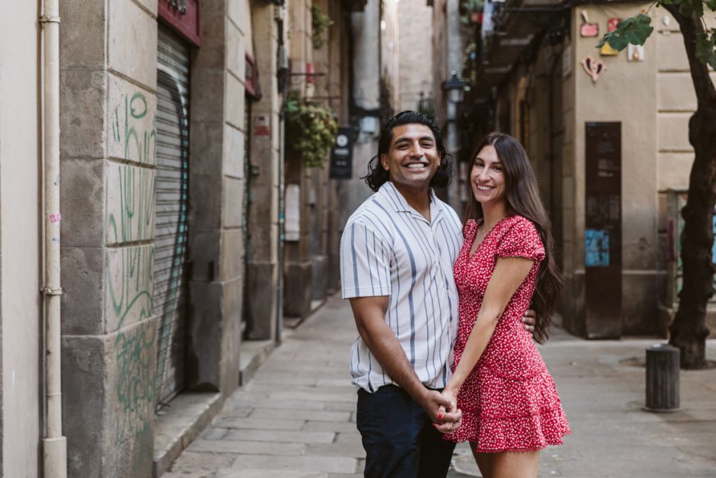 A smiling couple stands close together on a narrow city street. The man wears a white striped shirt and dark pants, while the woman wears a short red dress with white patterns. They hold hands and face the camera, surrounded by old buildings.