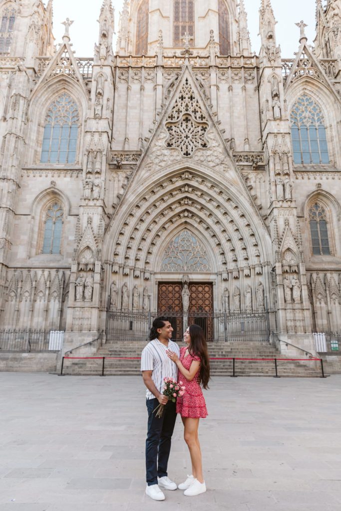 A couple stands facing each other in front of a large, ornate Gothic cathedral. The woman, in a red dress, holds a bouquet and touches the mans face; both are smiling. The cathedral’s detailed stone facade fills the background.