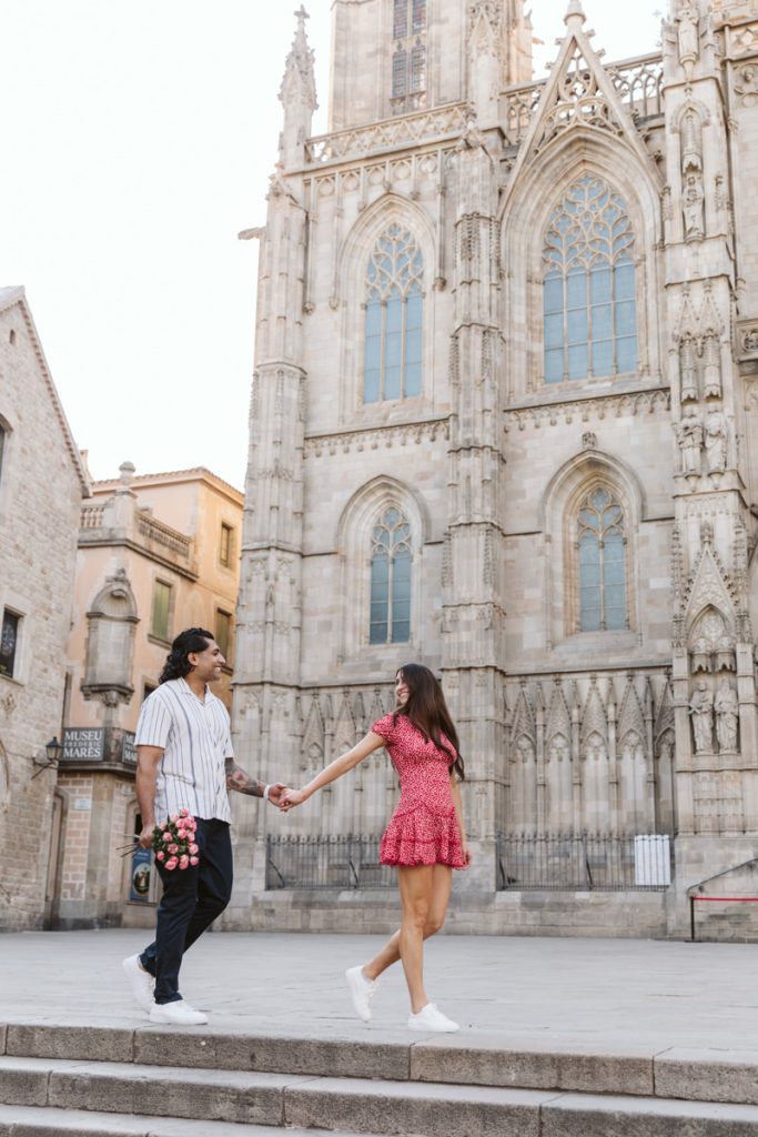 A couple holding hands walks in front of a historic Gothic-style cathedral. The man holds a bouquet of roses, and the woman wears a red dress. The scene is bright and romantic, with old stone buildings in the background.