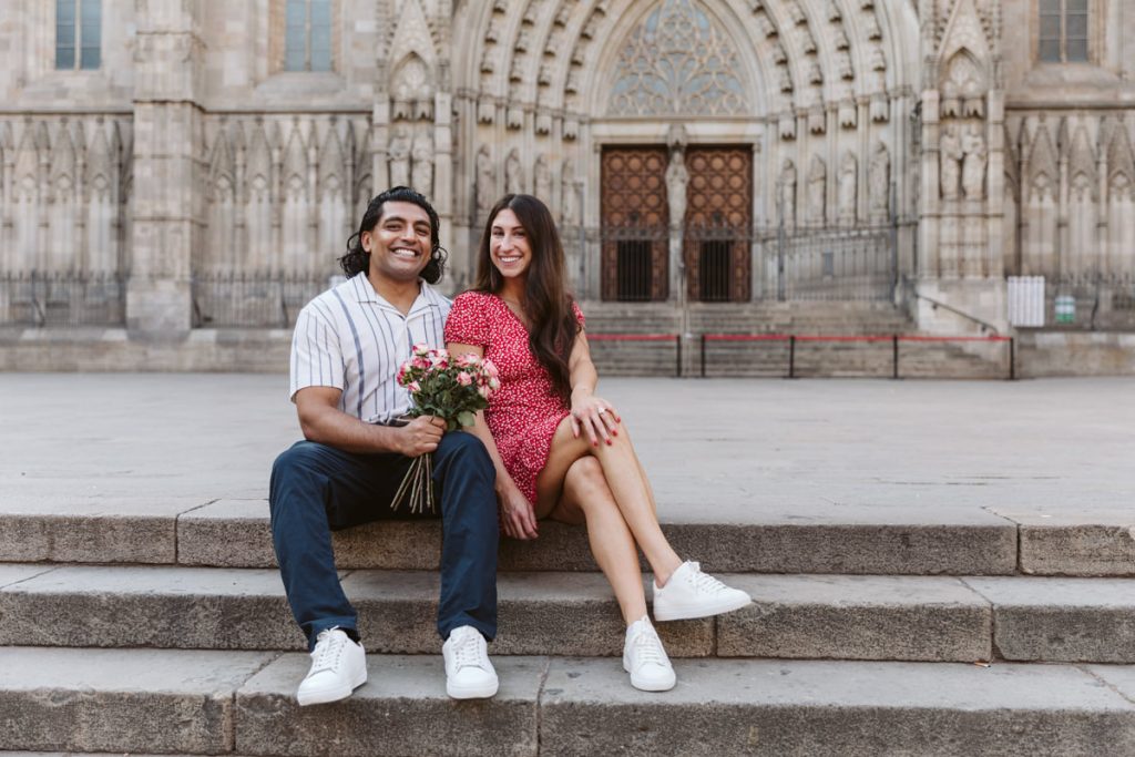 A smiling couple sits on stone steps in front of a large, ornate building. The man holds a bouquet of flowers, and the woman wears a red dress. Both appear happy and relaxed.