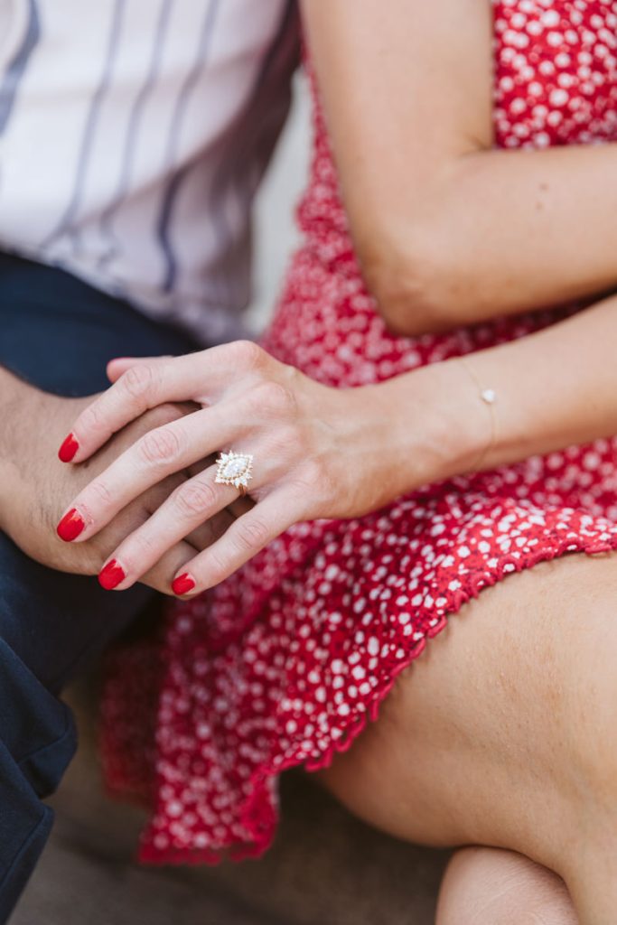 A close-up of a womans hand with red nail polish resting on her leg, wearing a large diamond ring. She is dressed in a red patterned dress, and another person is partially visible beside her.