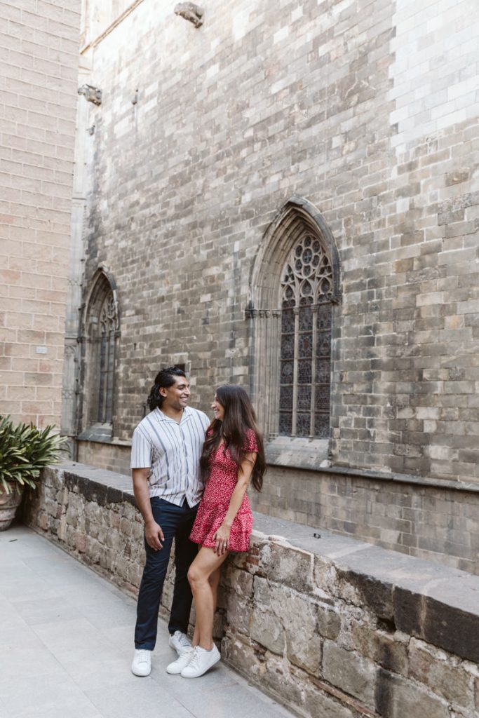 A couple stands and smiles at each other in front of a historic stone building with arched windows. The woman in a red dress sits on a low wall while the man, in a striped shirt, stands beside her.