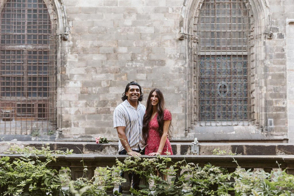 A smiling couple stands together in front of an old stone building with large, arched, barred windows. Greenery grows along the stone ledge in front of them. The woman wears a red dress; the man wears a striped shirt.
