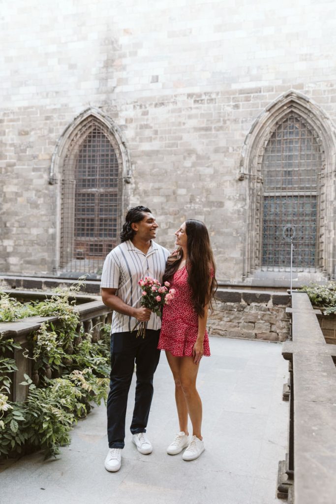 A couple stands outside in front of a stone building with arched windows. The man holds a bouquet of flowers, and they smile at each other. Both are casually dressed; greenery lines the walkway beside them.