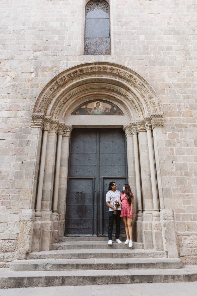 A couple stands on the steps in front of a large, old stone building with arched doorway and a painted religious icon above the entrance. They smile at each other, holding hands and flowers.