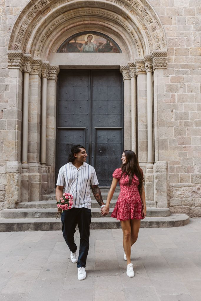 A couple holding hands and smiling at each other walks in front of a large historic stone building with an arched doorway. The man holds a bouquet of flowers behind his back, and both wear casual outfits with sneakers.