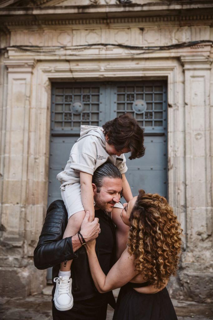 A man carries a young boy on his shoulders in front of an old stone building, while a curly-haired woman smiles warmly at them—capturing the joy and affection of family photos in the Gothic Quarter in Barcelona.