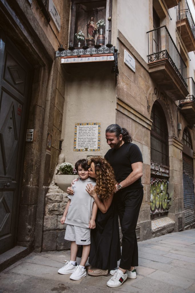 A woman kisses a young boy on the cheek as a man hugs them both. They stand on a narrow street beside an old building with a shrine and plaque—capturing the warmth of family photos in the Gothic Quarter in Barcelona.