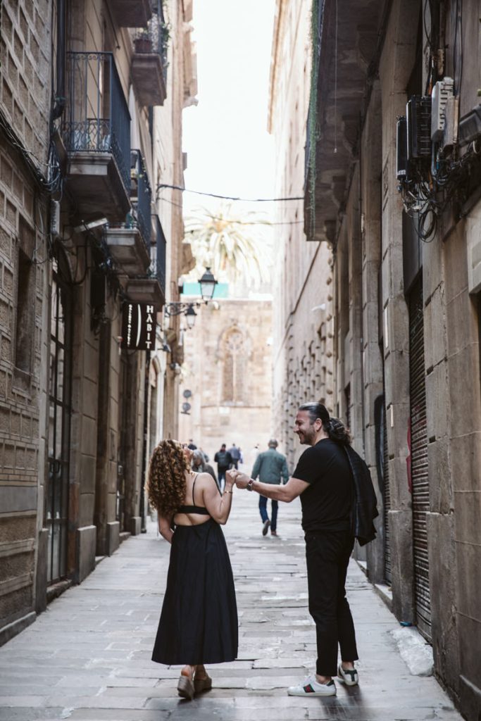 A couple walks hand-in-hand down a narrow, cobblestone street lined with old buildings in the Gothic Quarter in Barcelona—a perfect setting for timeless family photos, with a palm tree and others visible in the distance.