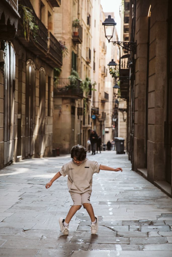 A young child in light-colored clothes joyfully skips along a narrow, sunlit cobblestone street in the Gothic Quarter in Barcelona, perfect for capturing family photos amid old buildings and charming balconies.
