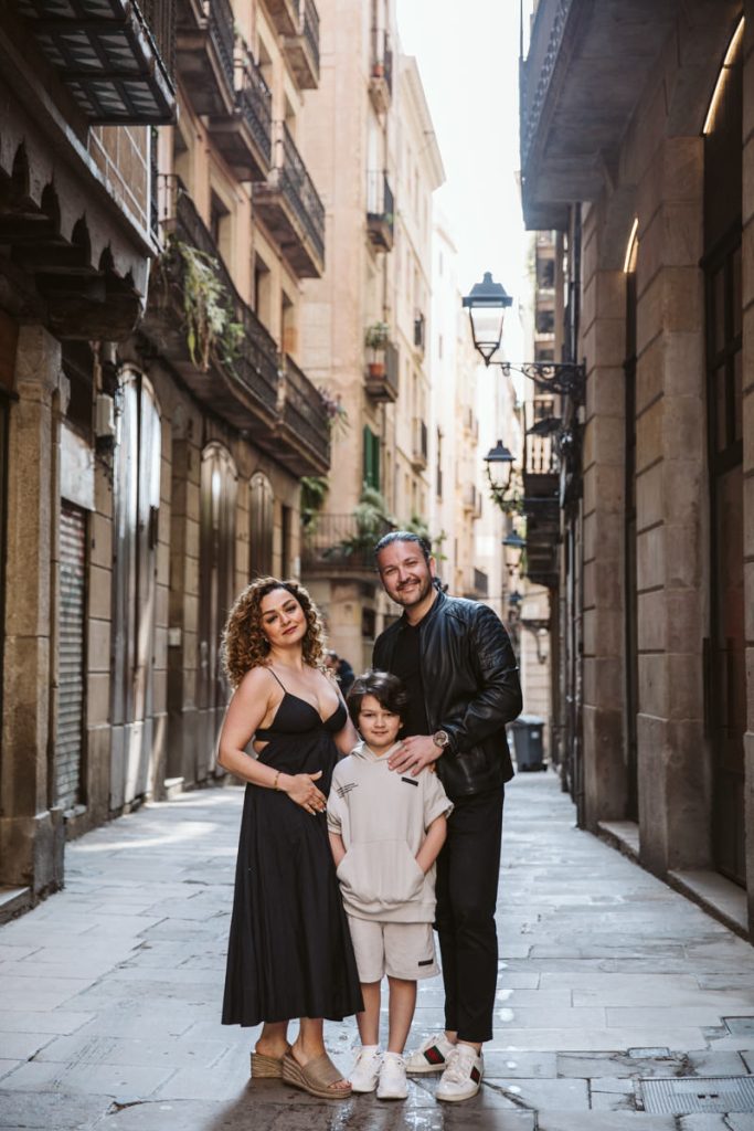 A family of three poses together on a narrow cobblestone street lined with old buildings, capturing family photos in the Gothic Quarter in Barcelona. The woman wears a black dress, the man is in black with a jacket, and the child wears light colors and white shoes.