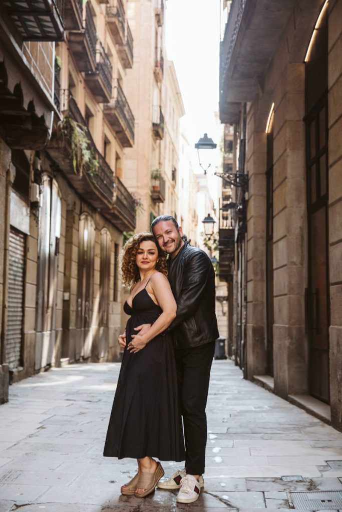 A couple poses closely together and smiles on a narrow, sunlit street lined with balconies—perfect for family photos in the Gothic Quarter in Barcelona. The woman wears a black dress and heels; the man stands behind her in a leather jacket and sneakers.