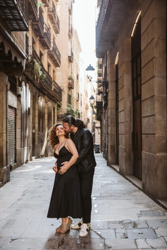 A couple stands in a narrow, sunlit street lined with old buildings in the Gothic Quarter of Barcelona. The man kisses the woman’s head from behind as she smiles, holding her baby bump—creating intimate family photos in stylish black outfits.