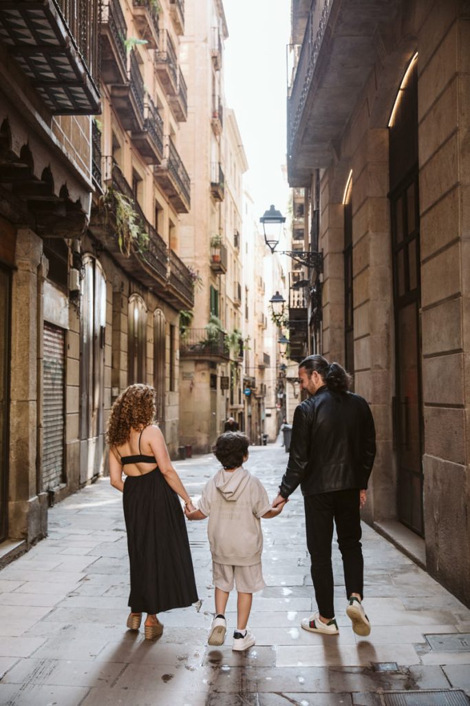A family strolls hand in hand down a narrow, old city street lined with balconies and tall buildings—perfect for family photos in the Gothic Quarter in Barcelona.