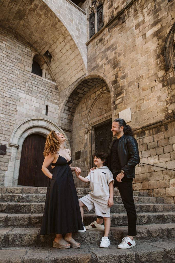 A woman in a black dress, a man in a black jacket, and a child in light clothing smile together on stone steps outside a historic building—perfect for family photos in the Gothic Quarter in Barcelona.
