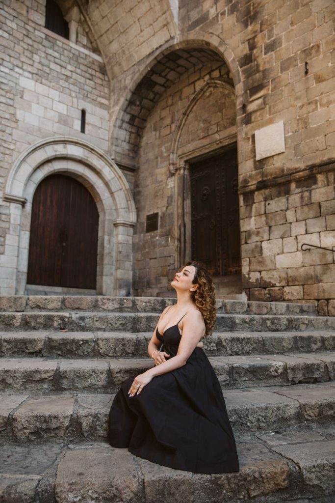A woman in a long black dress kneels on old stone steps in front of an arched doorway and historic stone building—evoking the timeless charm of family photos in the Gothic Quarter in Barcelona, eyes closed and face tilted upward.