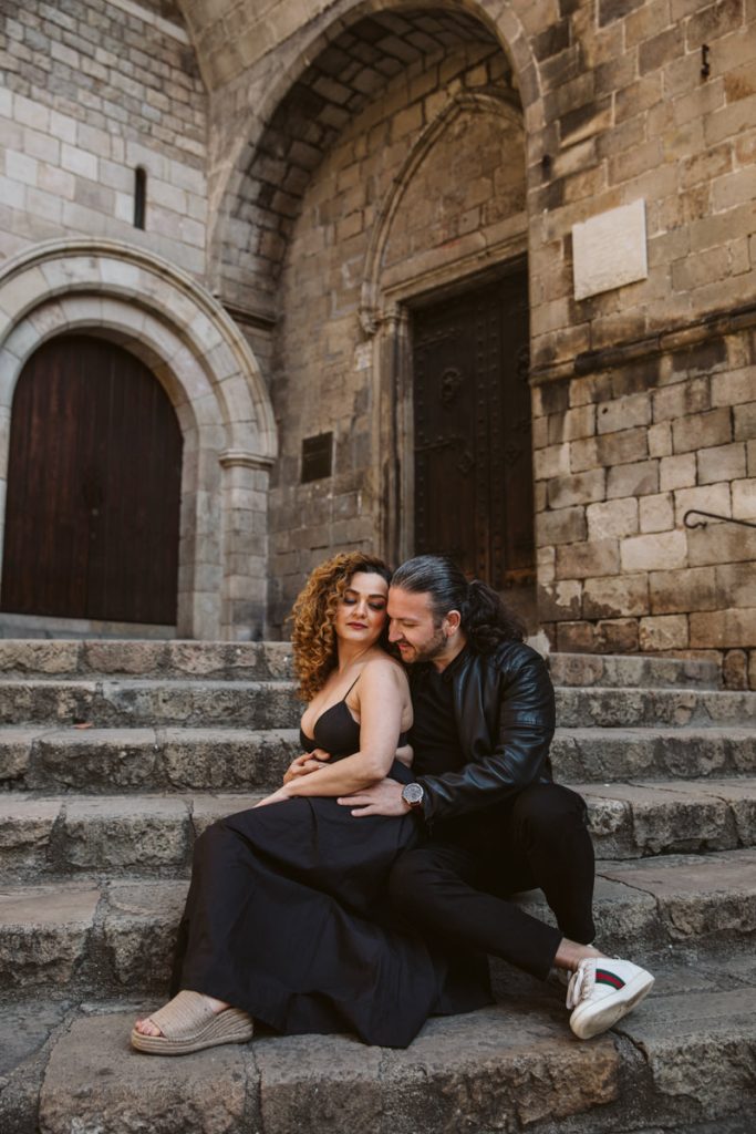 A couple sits closely together on stone steps in front of an old brick building, capturing the romance often seen in family photos in the Gothic Quarter in Barcelona. She wears a black dress and sandals, while he lovingly embraces her from behind.