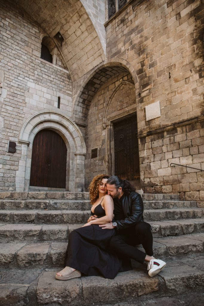 A couple sits closely on stone steps in front of an old building with arched wooden doors, capturing a tender moment reminiscent of family photos in the Gothic Quarter in Barcelona. The woman in a black dress leans into the man, who embraces her affectionately.