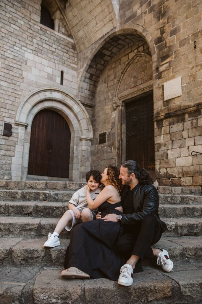 A family of three sits on stone steps outside an old stone building, capturing family photos in the Gothic Quarter in Barcelona. The mother hugs and kisses their smiling child while the father sits beside them, all appearing happy and relaxed.