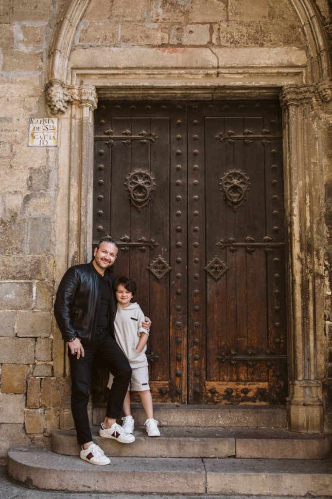 A man and a boy stand smiling together in front of a large, ornate wooden door set in an old stone building—perfect for family photos in the Gothic Quarter in Barcelona. The boy hugs the man, and both wear casual clothes and sneakers.