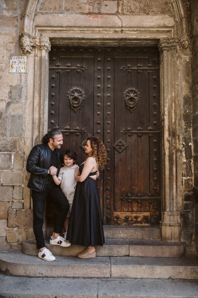 A man, woman, and child stand smiling together on stone steps in front of a large, ornate wooden door with stone arches—capturing family photos in the Gothic Quarter in Barcelona.
