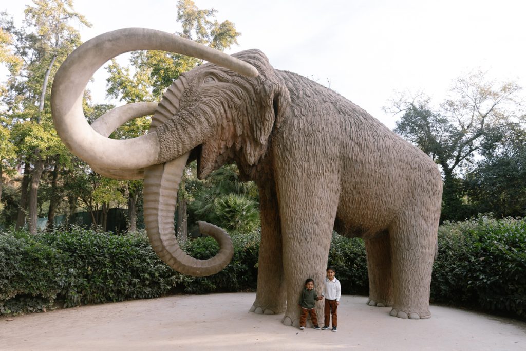 Two children stand beneath a large, realistic mammoth statue with long curved tusks in an outdoor park surrounded by trees and greenery.