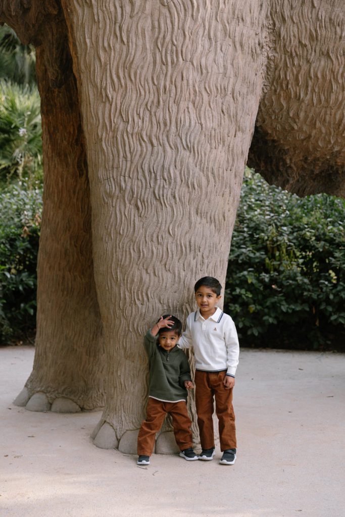 Two young boys stand beside the large leg of a statue resembling an elephant, with greenery in the background. One boy touches the statues leg while both smile at the camera.