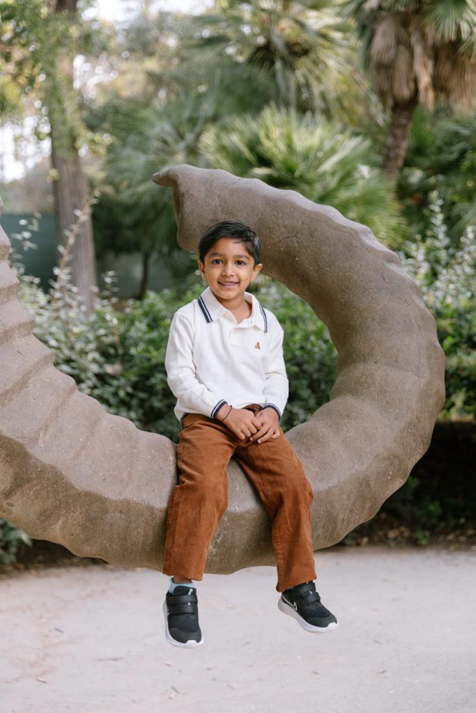 A young boy with short dark hair, wearing a white long-sleeve shirt and brown pants, sits and smiles on a large, curved stone sculpture in an outdoor garden setting.