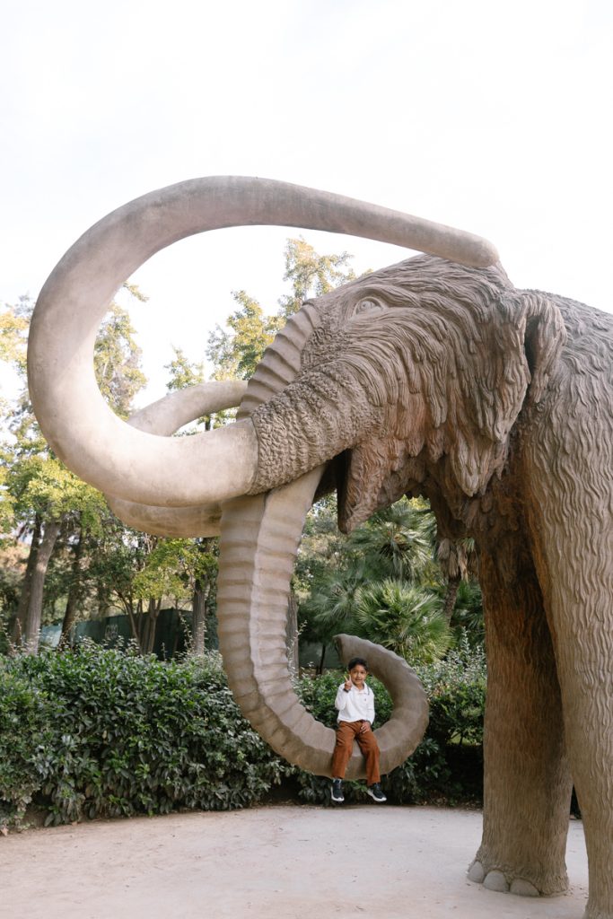 A person sits on the curled trunk of a large woolly mammoth statue in an outdoor park, surrounded by greenery and trees.
