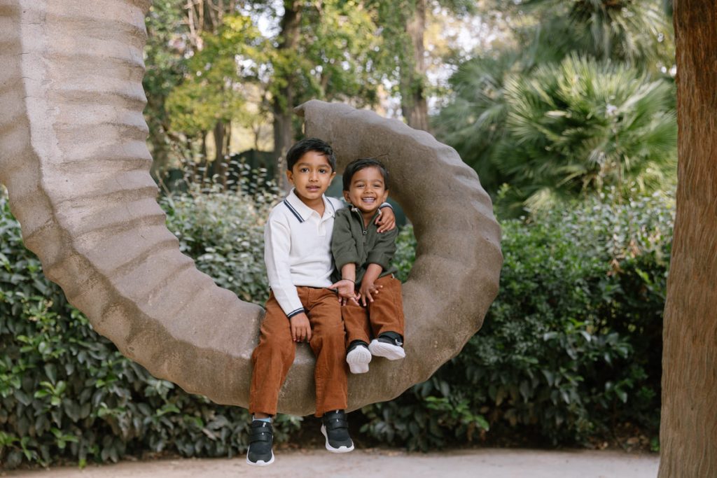 Two young boys sit together on a large, curved stone sculpture outdoors, surrounded by green foliage. One boy is smiling widely and hugging the other, who has a calm expression. Both wear brown pants and sneakers.