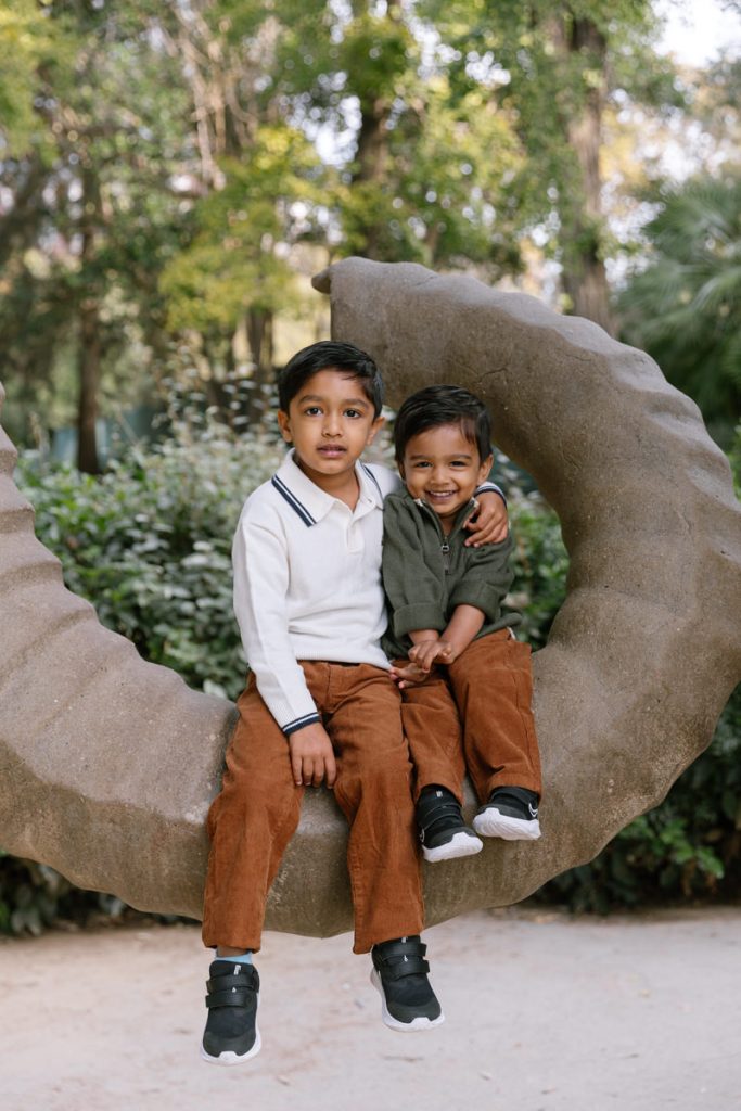 Two young boys sit closely together on a large, curved stone sculpture outdoors. They are smiling, wearing brown pants and sneakers, with trees and greenery in the background.