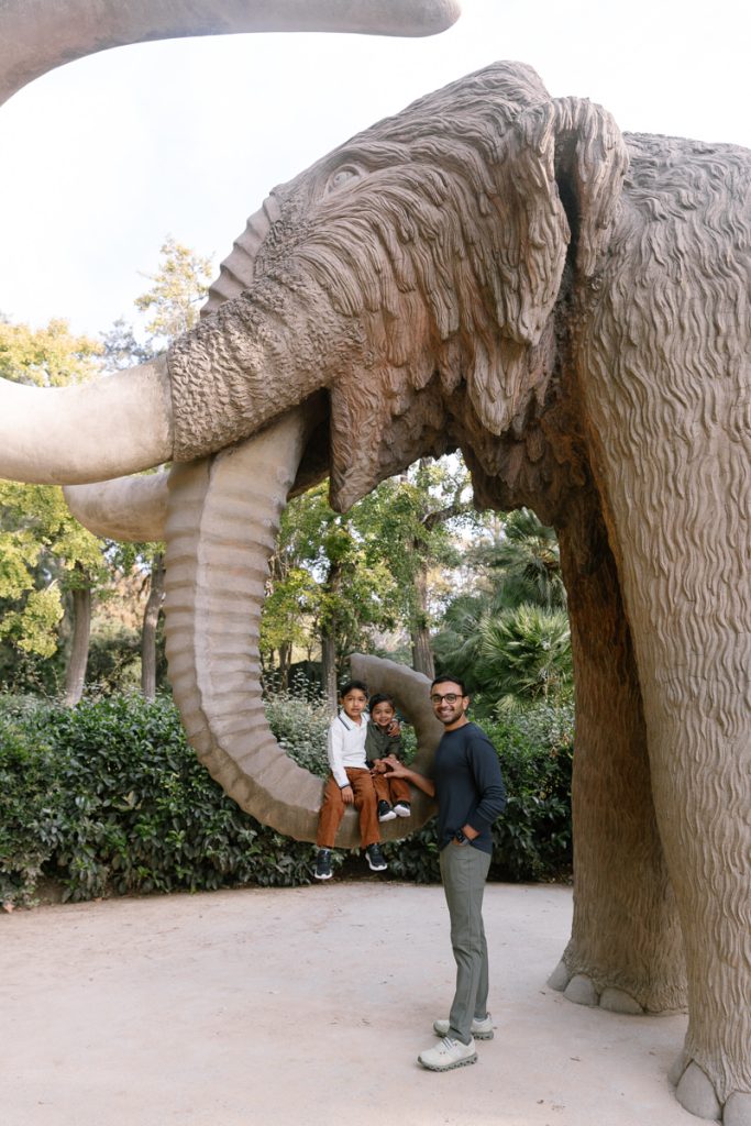 A man stands smiling near a large mammoth statue outdoors, while two children sit together on one of the mammoths tusks, surrounded by greenery and trees.