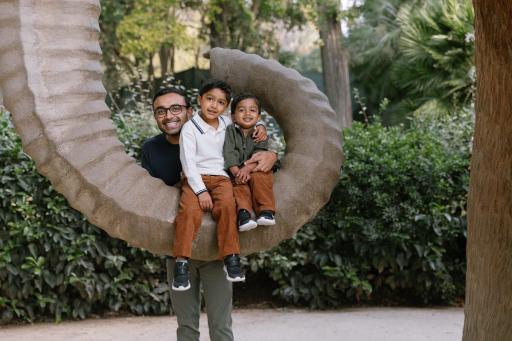 A man and two young boys sit together on a large, curved stone sculpture outdoors, smiling at the camera. Lush green plants and trees fill the background.
