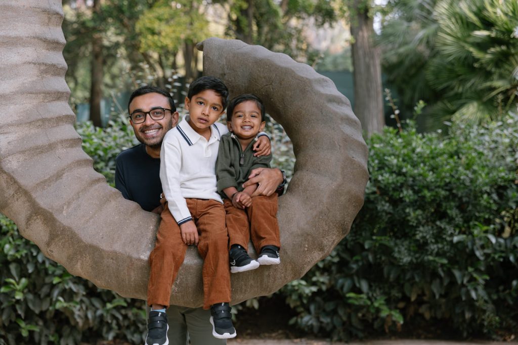 A man smiles with two young boys sitting on a large curved stone sculpture in an outdoor park, surrounded by greenery and trees.