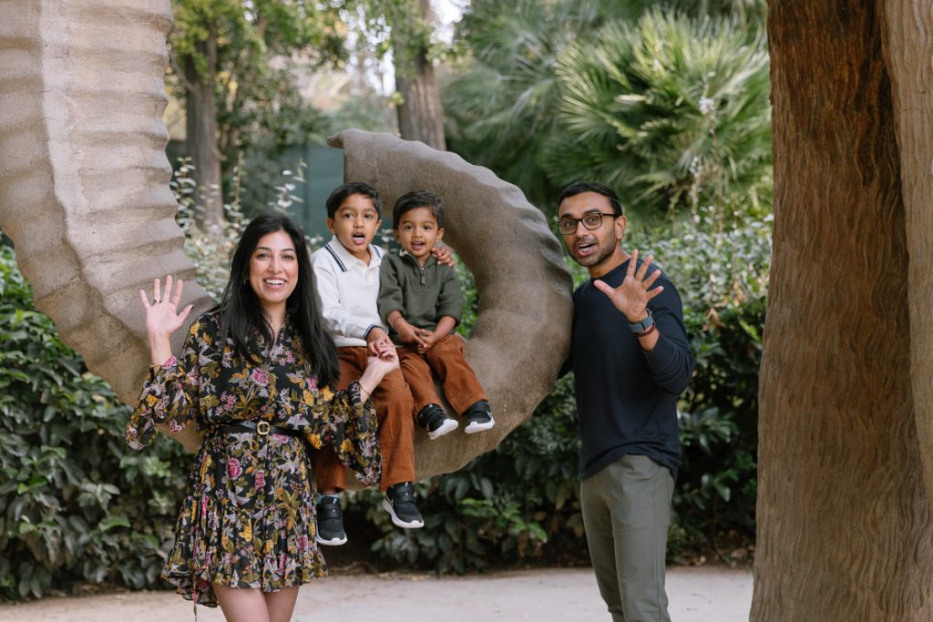 A smiling family of four waves at the camera outdoors. Two children sit on a large, curved sculpture, while their parents stand on either side. Trees and greenery fill the background.