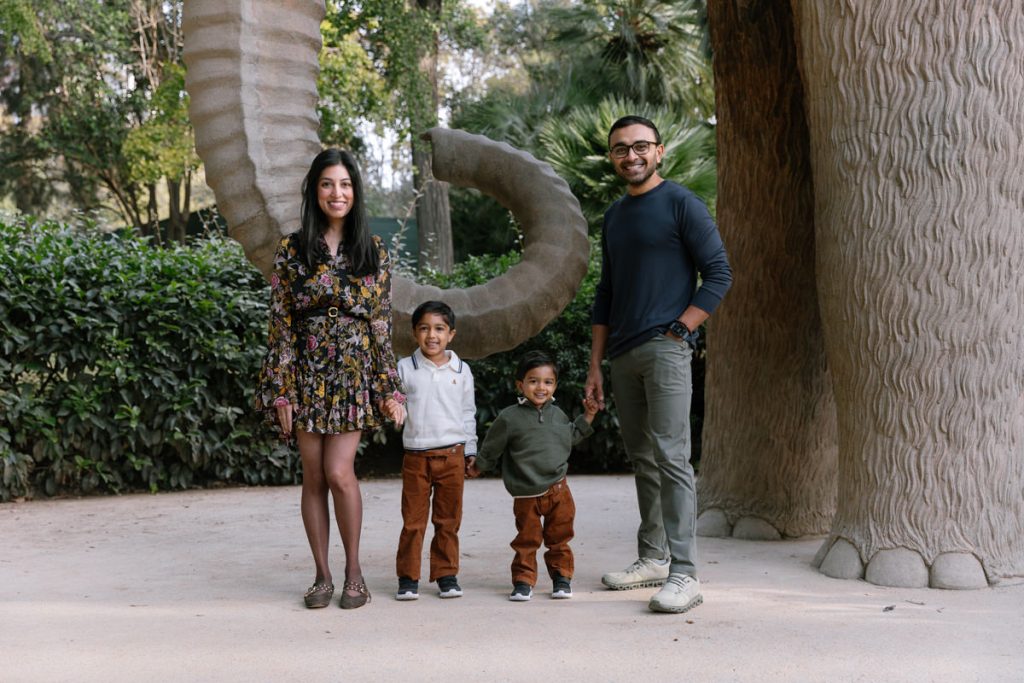 A family of four poses outdoors near large faux elephant legs and trunk. The woman wears a floral dress, the man a dark shirt and jeans, and two young boys stand between them, all smiling. Lush greenery is in the background.