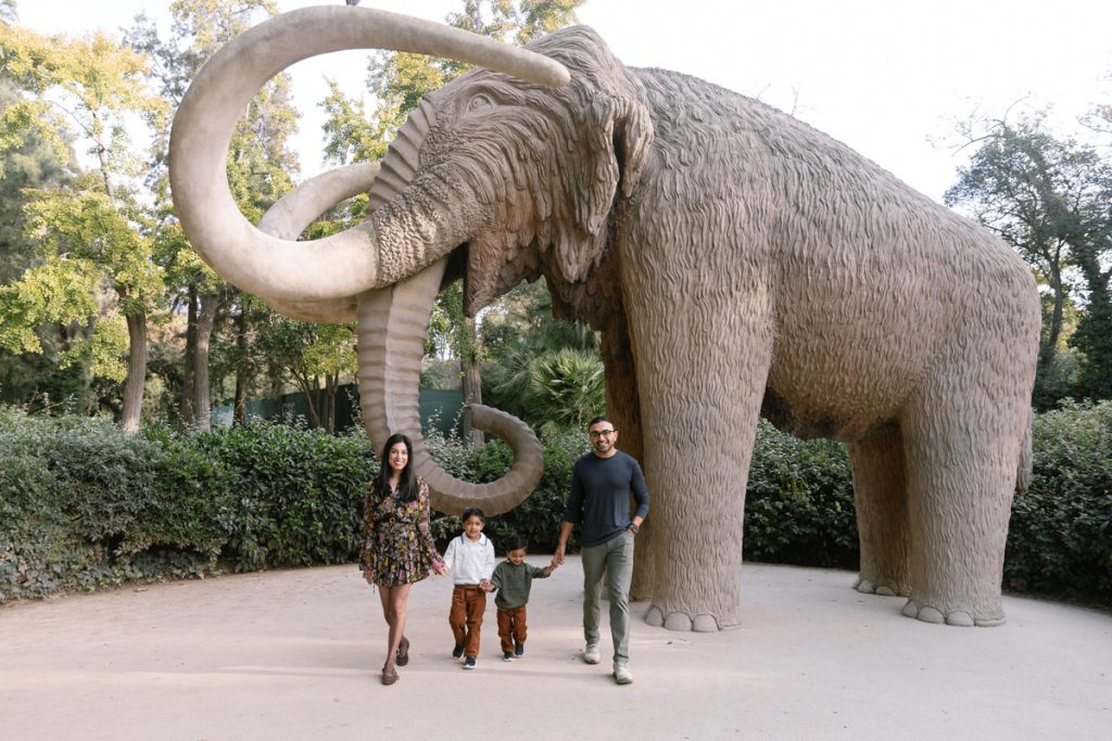A family of four, two adults and two children, pose and hold hands in front of a large woolly mammoth statue in a park surrounded by greenery.