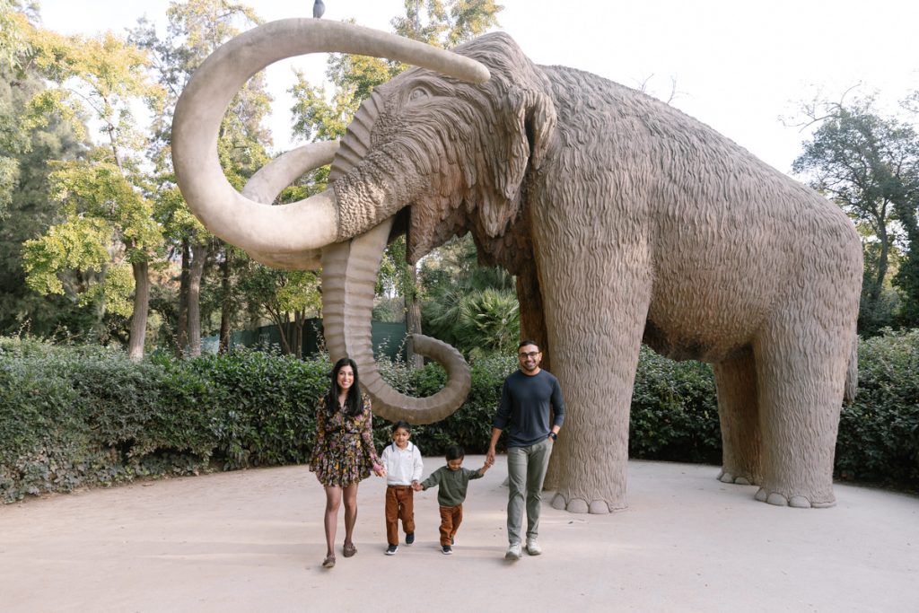 A family of four stands in front of a large woolly mammoth statue in a park, surrounded by greenery and trees. The adults and children are smiling and holding hands.