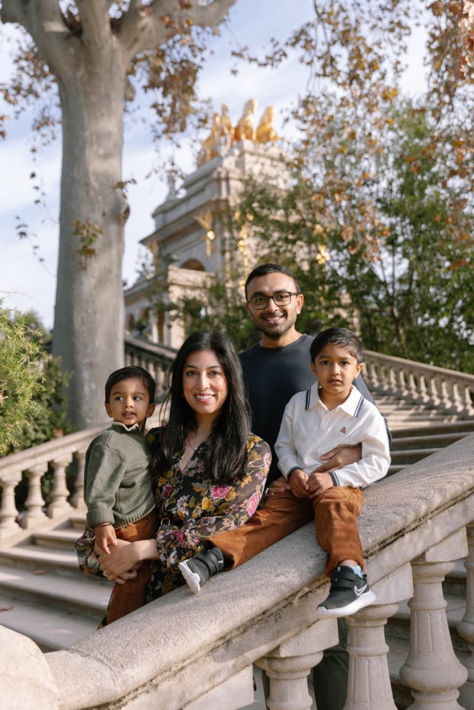 A family of four poses on outdoor stone steps. Two adults and two young children smile at the camera, surrounded by trees and an ornate building in the background. The scene is bright and warm.