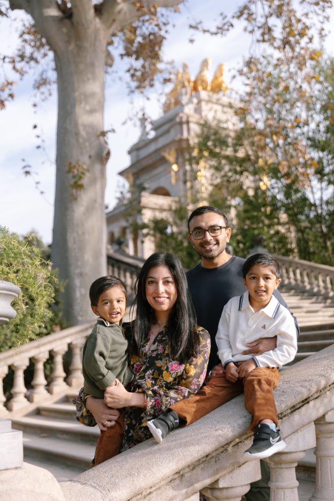 A smiling family of four poses on outdoor stone steps, with trees and a decorative building in the background. The mother holds a young child, while the father stands behind her next to an older child sitting on the railing.