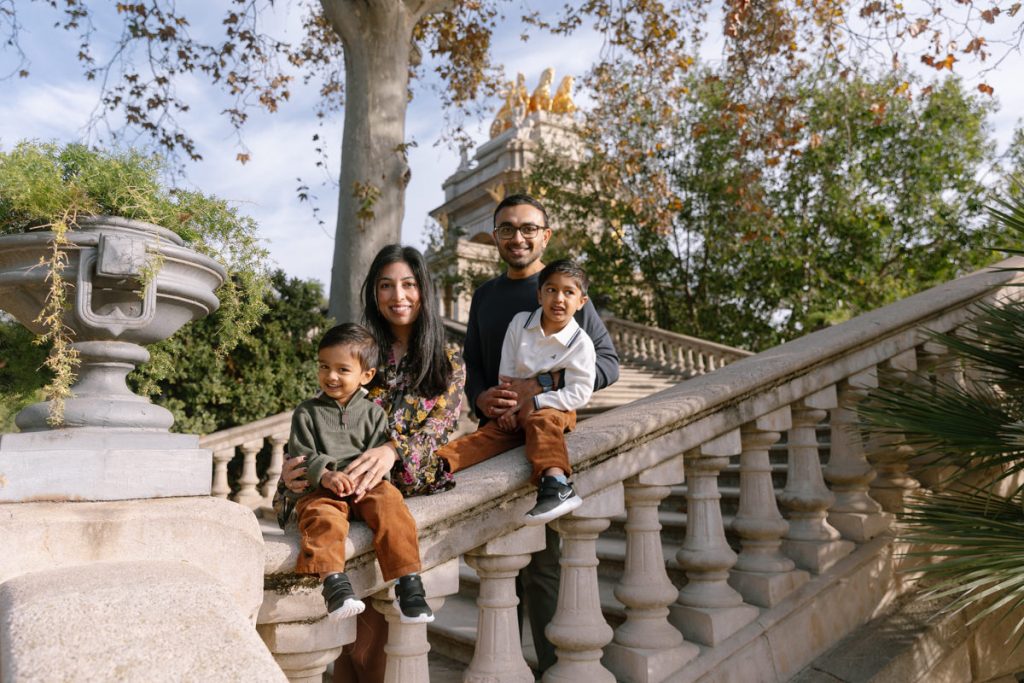 A family of four poses on outdoor stone steps surrounded by greenery. The parents sit beside their two young children, all smiling and dressed in casual clothes on a sunny day.