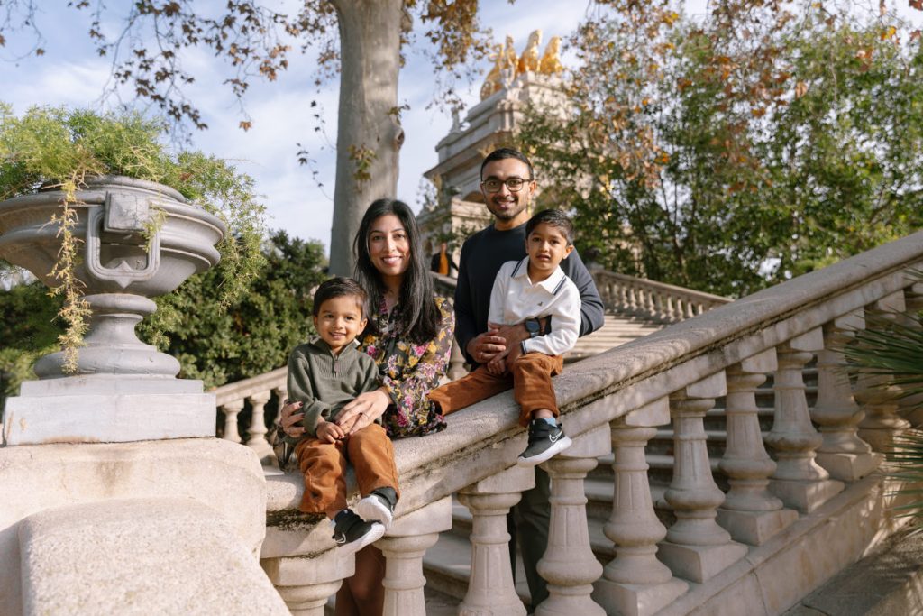 A family of four poses on a stone staircase outdoors, surrounded by trees. The parents sit close together, smiling, with their two young sons in front, all dressed in coordinated outfits.
