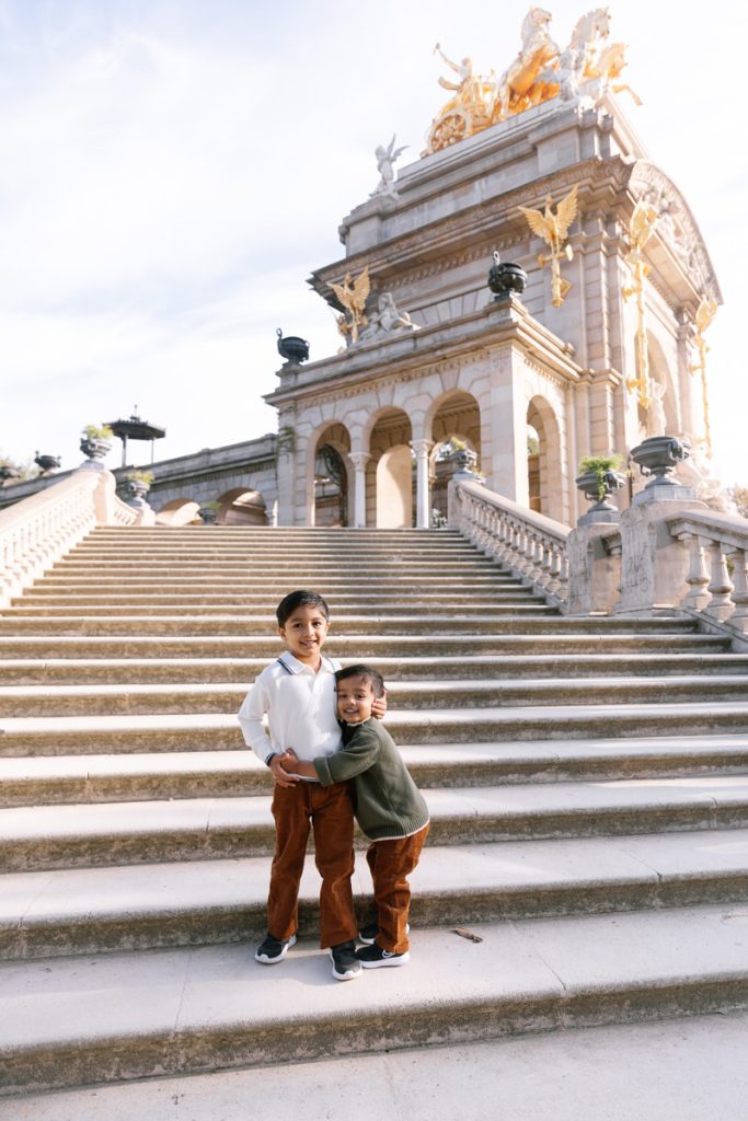 Two young children pose together, smiling on wide stone steps in front of a grand historic monument adorned with gold statues and intricate architectural details under a bright sky.