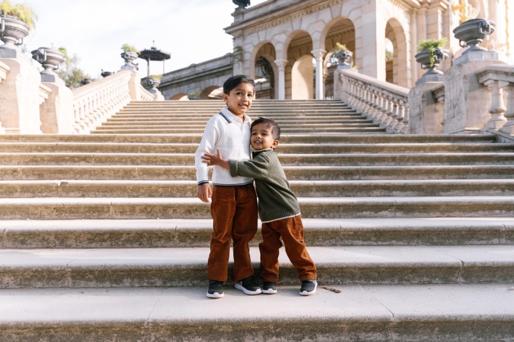 Two young children stand on wide stone steps outdoors. One child hugs the other, both smiling, with a grand building and stone railings in the background. They wear casual sweaters and brown pants.