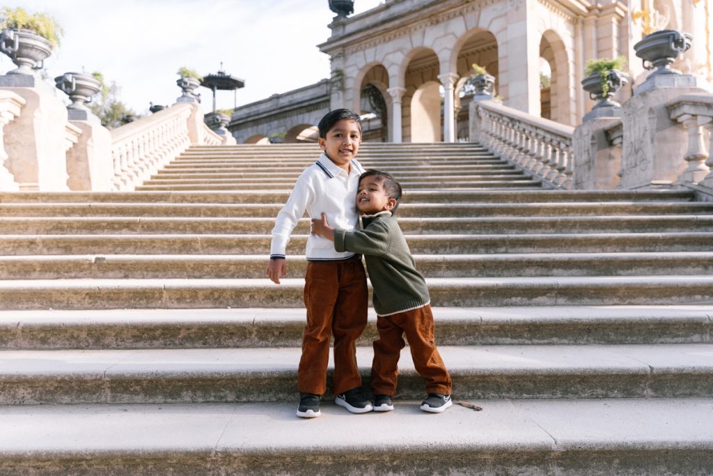 Two young boys stand on wide stone steps outdoors, smiling and hugging. They wear brown pants and sweaters, with an elegant historic building and balustrades in the background.