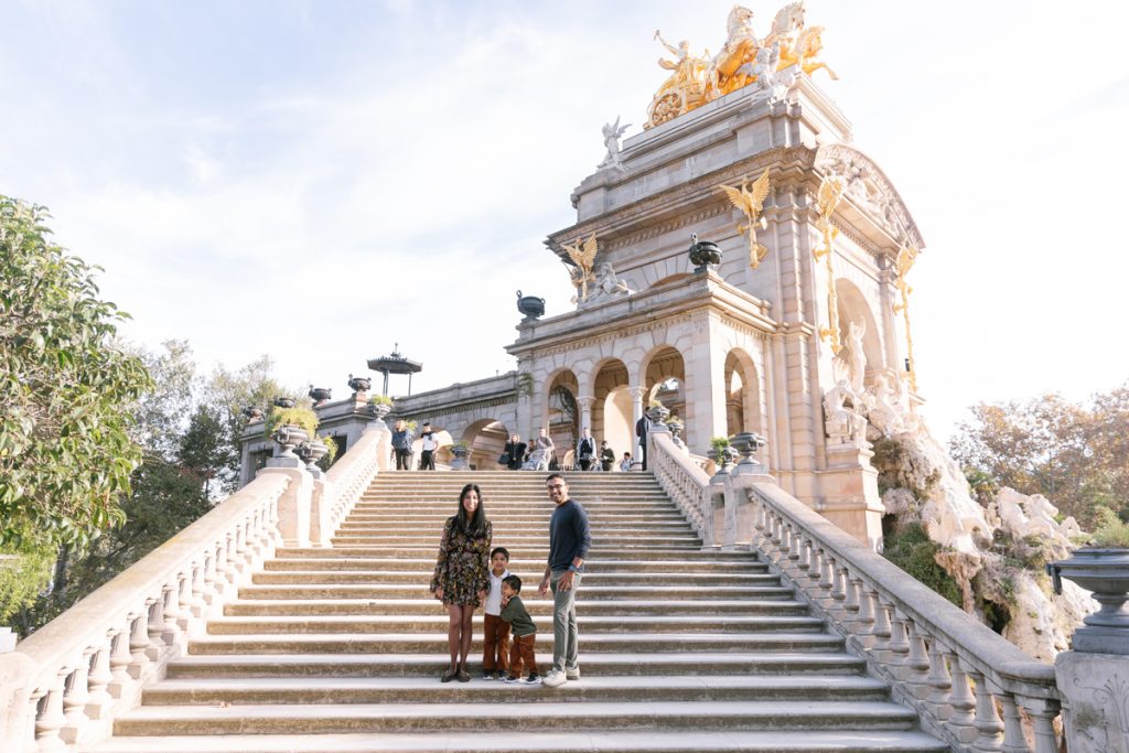A family of four stands on the steps of the ornate Cascada Monumental fountain in Barcelona’s Parc de la Ciutadella, with bright sunlight illuminating the grand, golden sculpture atop the structure.