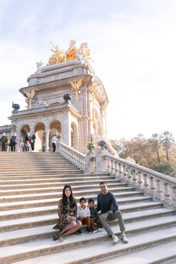 A family of four sits and smiles on wide stone steps in front of a grand, ornate monument with golden sculptures on top, under a partly cloudy sky. Other people stand on the steps above them.