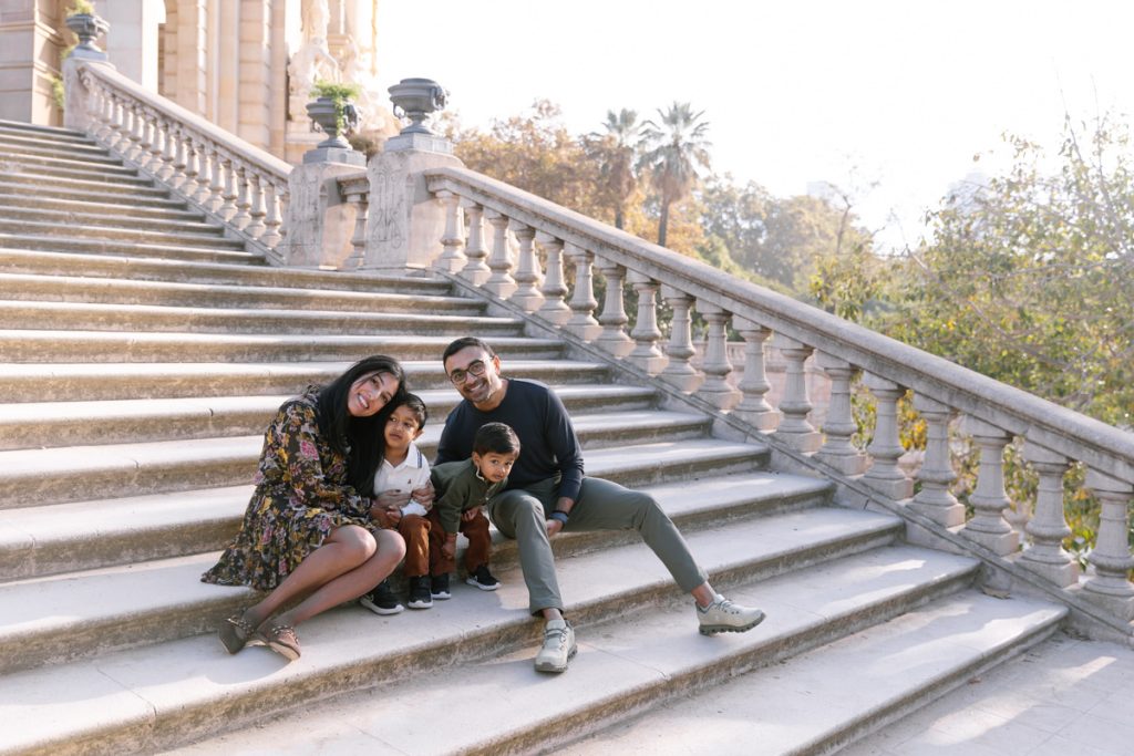 A family of four sits together on wide stone steps outdoors, smiling at the camera. Trees and ornate railings are in the background, suggesting a park or historic building setting.